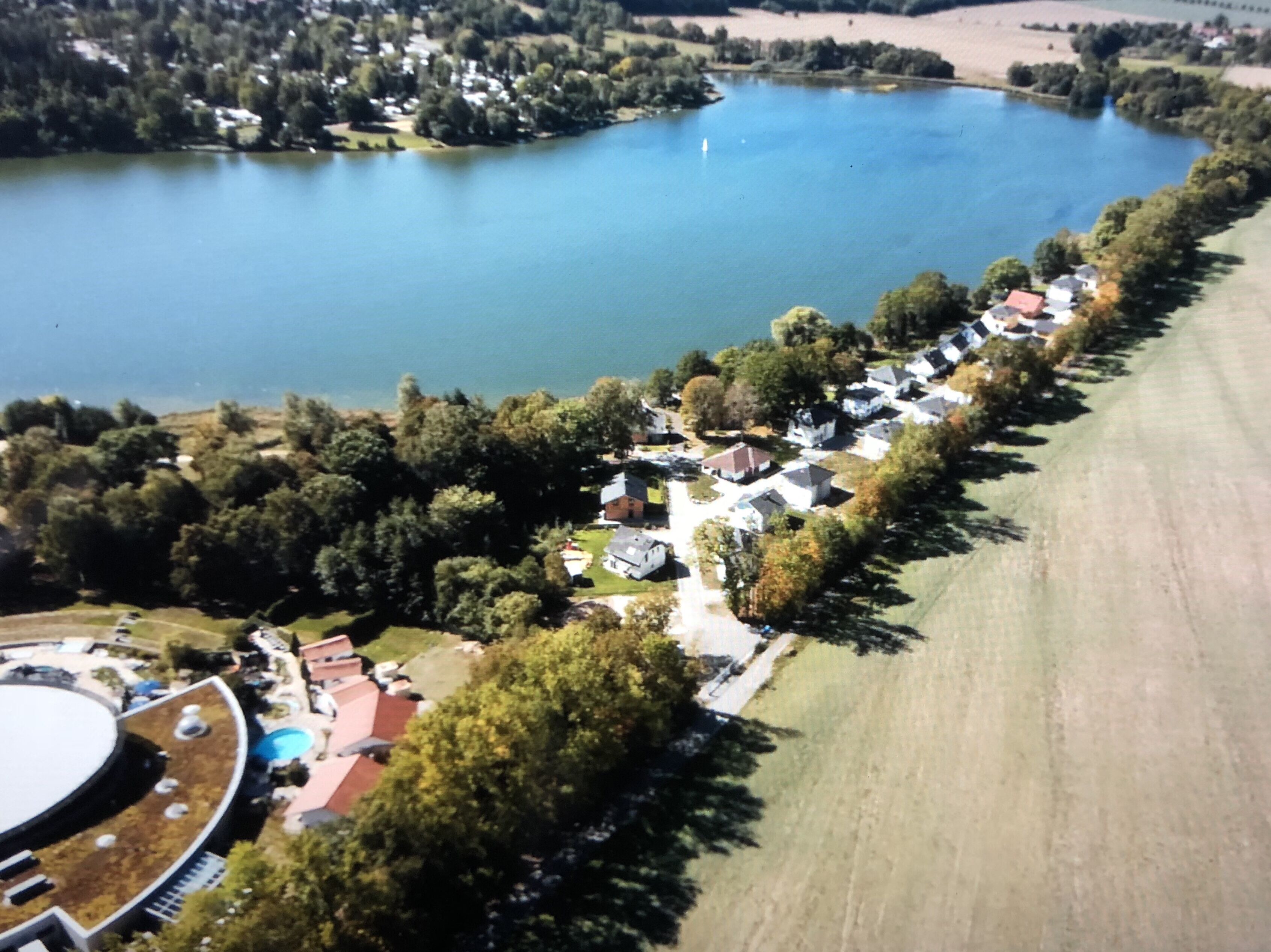 Vista aérea de uma área residencial à beira de um lago, com casas, piscina e um grande campo aberto.