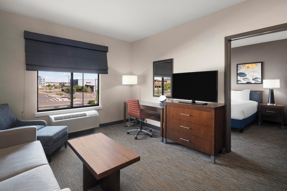Ein Hotelzimmer mit einer Couch, einem Schreibtisch, einem Fernseher, einem Bett, einer Kommode und einem Fenster mit Blick auf Gebäude.