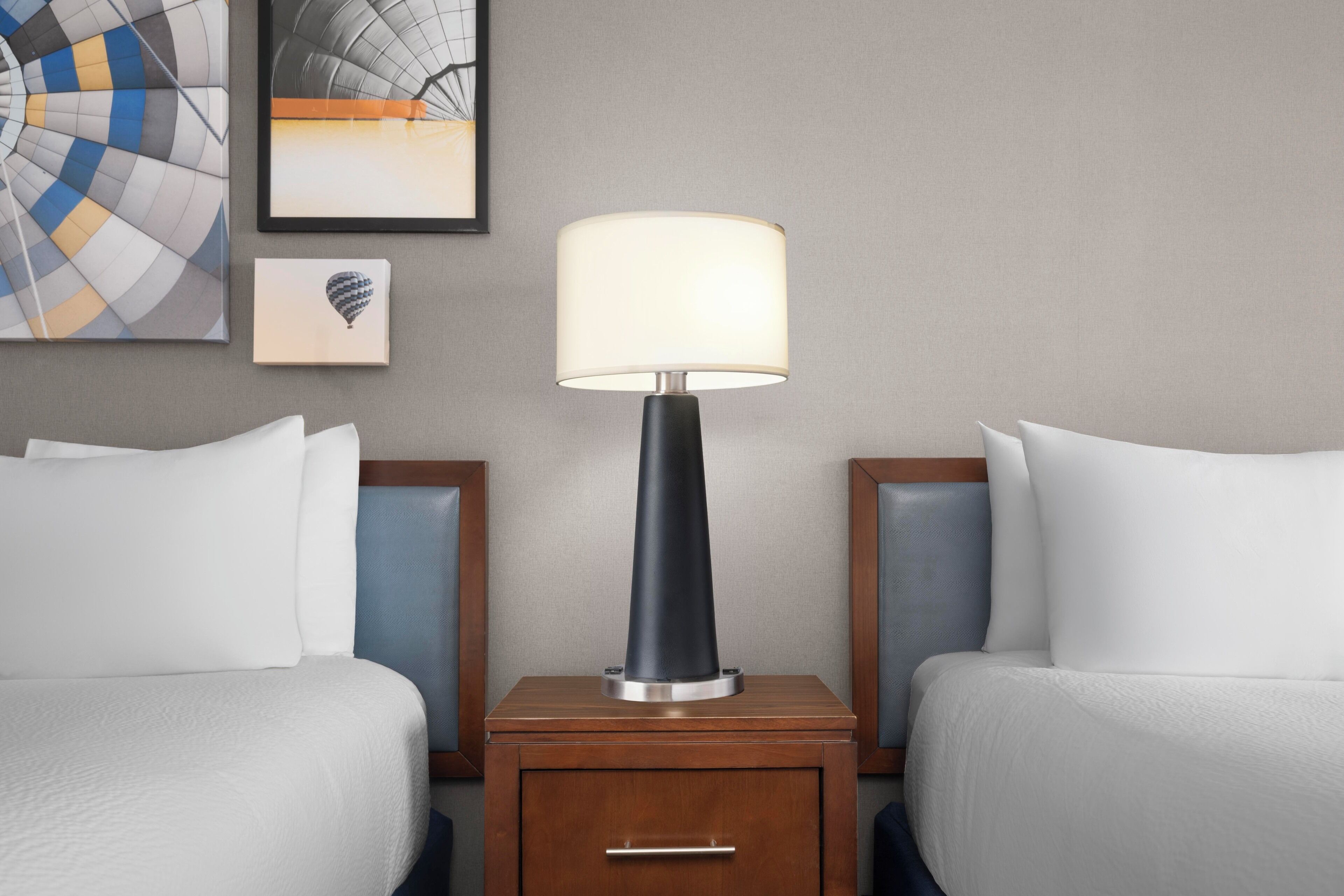 A hotel room with two beds, a nightstand with a lamp, and artwork on the wall.