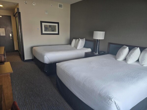 Habitación de hotel con dos camas, una mesita de noche con lámpara, un cuadro en la pared y piso alfombrado.