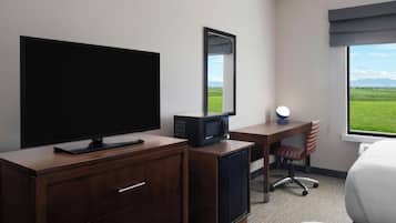 A hotel room with a TV, a desk with a chair, a small refrigerator, a mirror, and a window with a view of the outdoors.