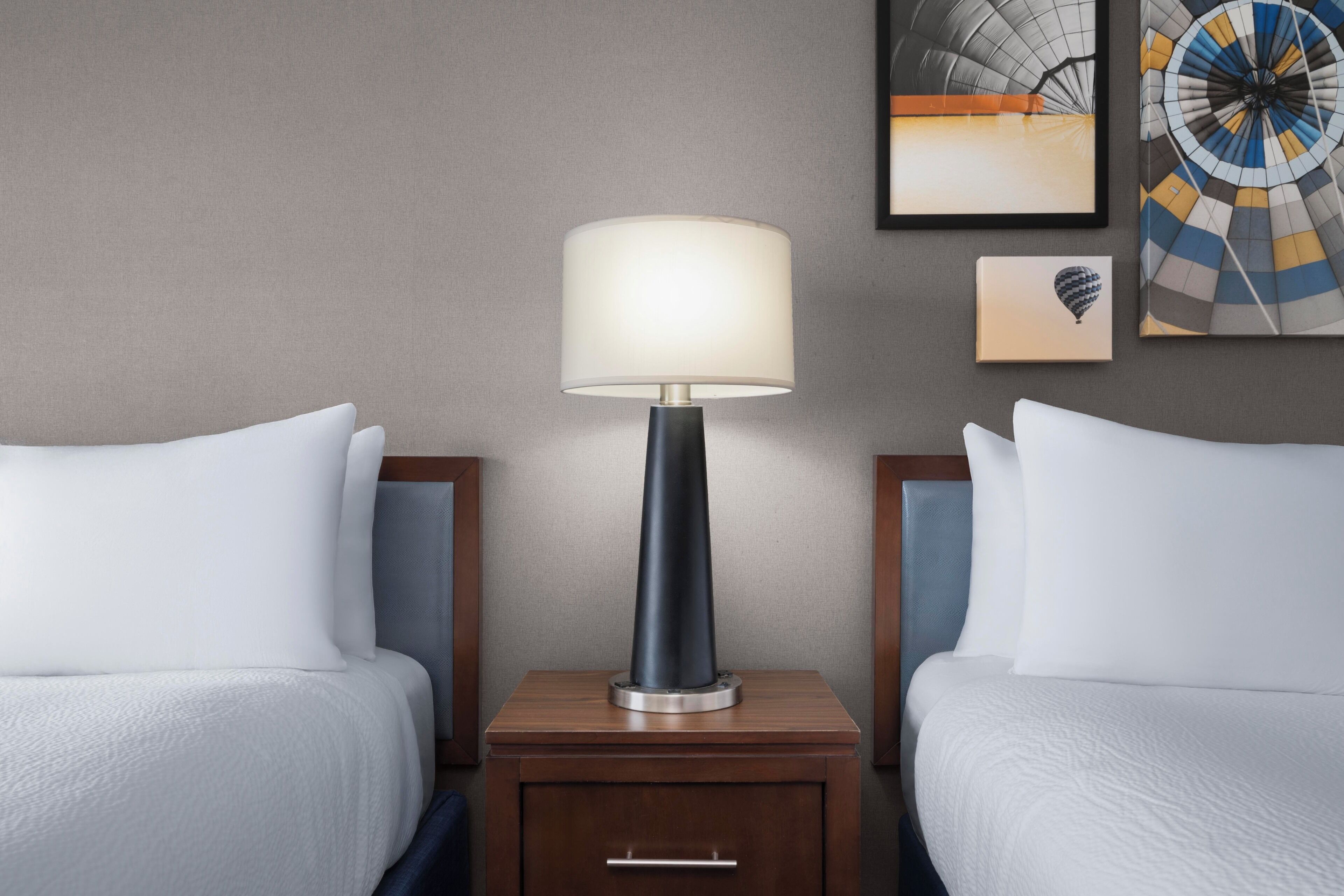 Een hotelkamer met twee bedden, een nachtkastje met een lamp, een klein ladeblok en twee ingelijste foto’s aan de muur.