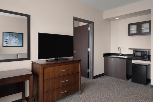 A hotel room with a TV, wooden dresser, kitchenette, and a framed picture on the wall.