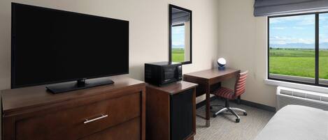 A hotel room with a flat-screen TV, a desk with a chair, a large window with a view of the outdoors, and a bed.