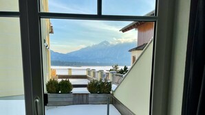A view from a window of a building with a balcony, snow-covered ground, and a mountain in the background. - 5 star apartment on Lake Hopfensee directly on the lake with 2 terraces & underground parking space (Hopfen am See)