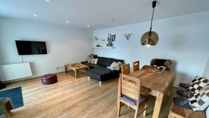 A modern living room with a wooden floor, a black sofa, a wooden dining table with chairs, a TV mounted on the wall, and a ceiling light fixture. - 5 star apartment on Lake Hopfensee directly on the lake with 2 terraces & underground parking space (Hopfen am See)