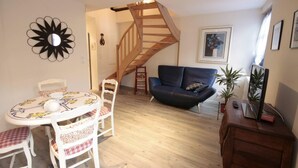 Un salon avec un escalier en bois, une table de salle à manger ronde entourée de chaises, un canapé et un meuble pour la télévision agrémenté d’une plante.