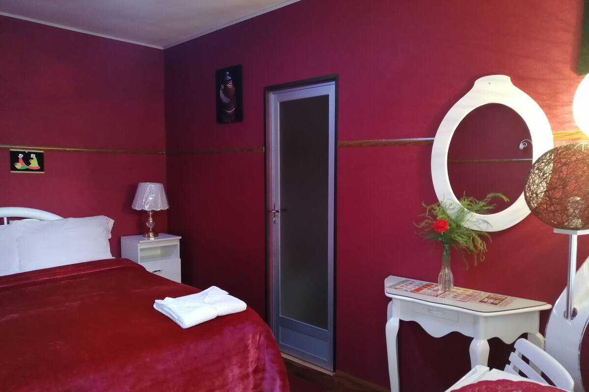Ein Hotelzimmer mit roter Bettdecke, einem weißen Nachttisch mit Lampe, einem Spiegel und einem kleinen Tisch mit Blumen.