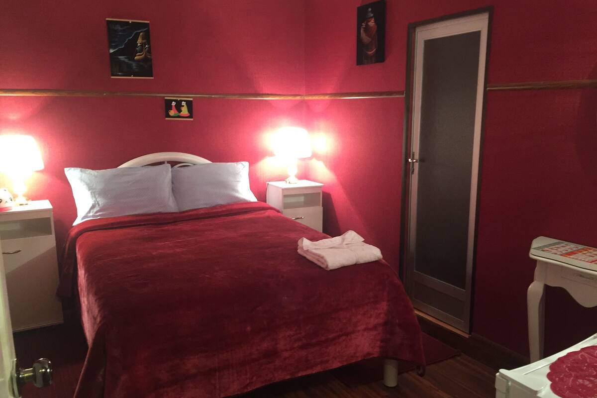 Ein Zimmer mit einem Bett, zwei Nachttischen, einem kleinen Tisch mit roter Decke und einer Tür.