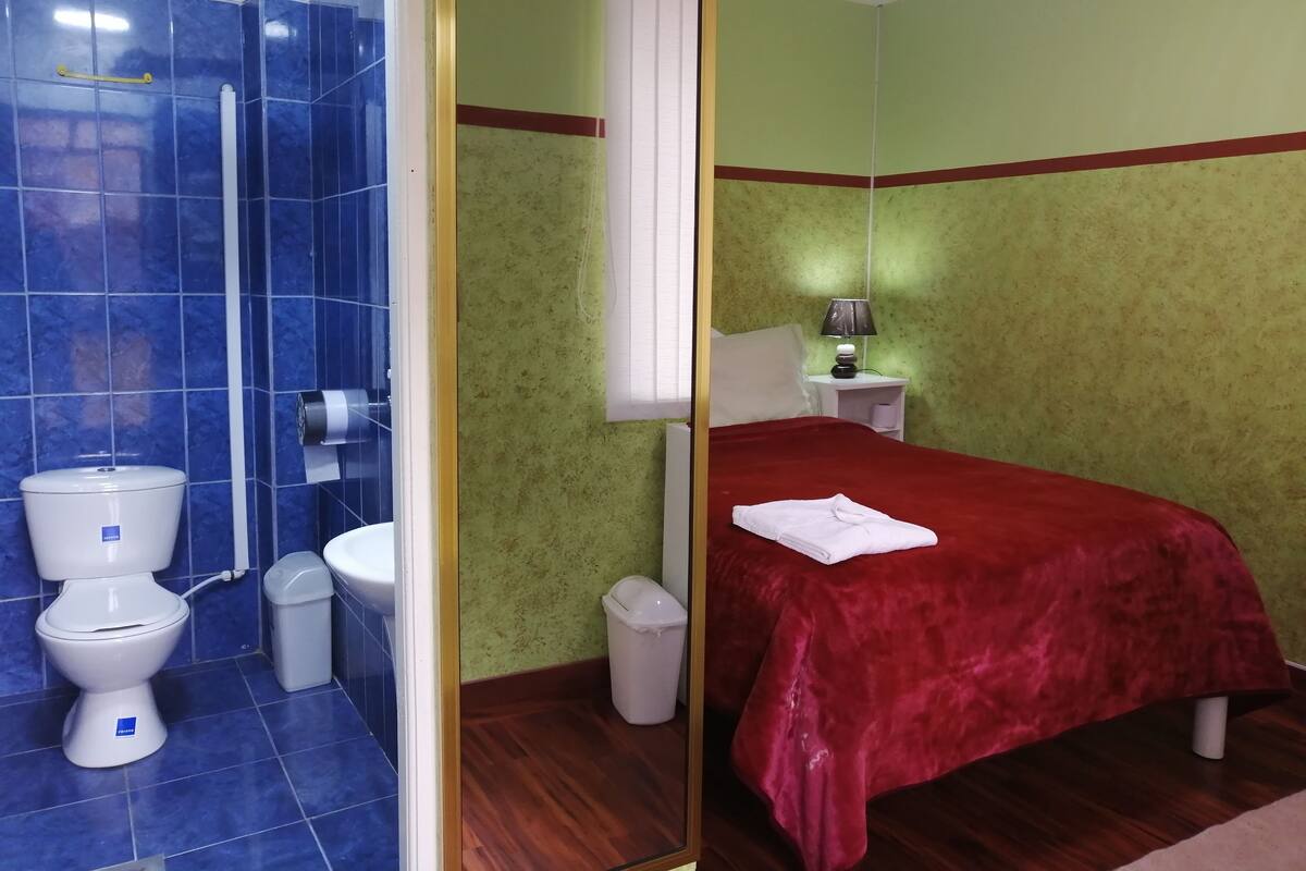 Ein Hotelzimmer mit Bad, das Toilette, Waschbecken und Dusche umfasst. Das Zimmer verfügt über ein Bett mit roter Bettdecke und einen Nachttisch mit Lampe.