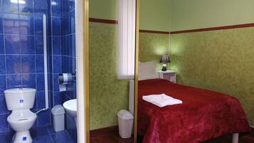 A hotel room with a bathroom featuring a toilet, sink, and shower. The room has a bed with a red bedspread and a nightstand with a lamp.