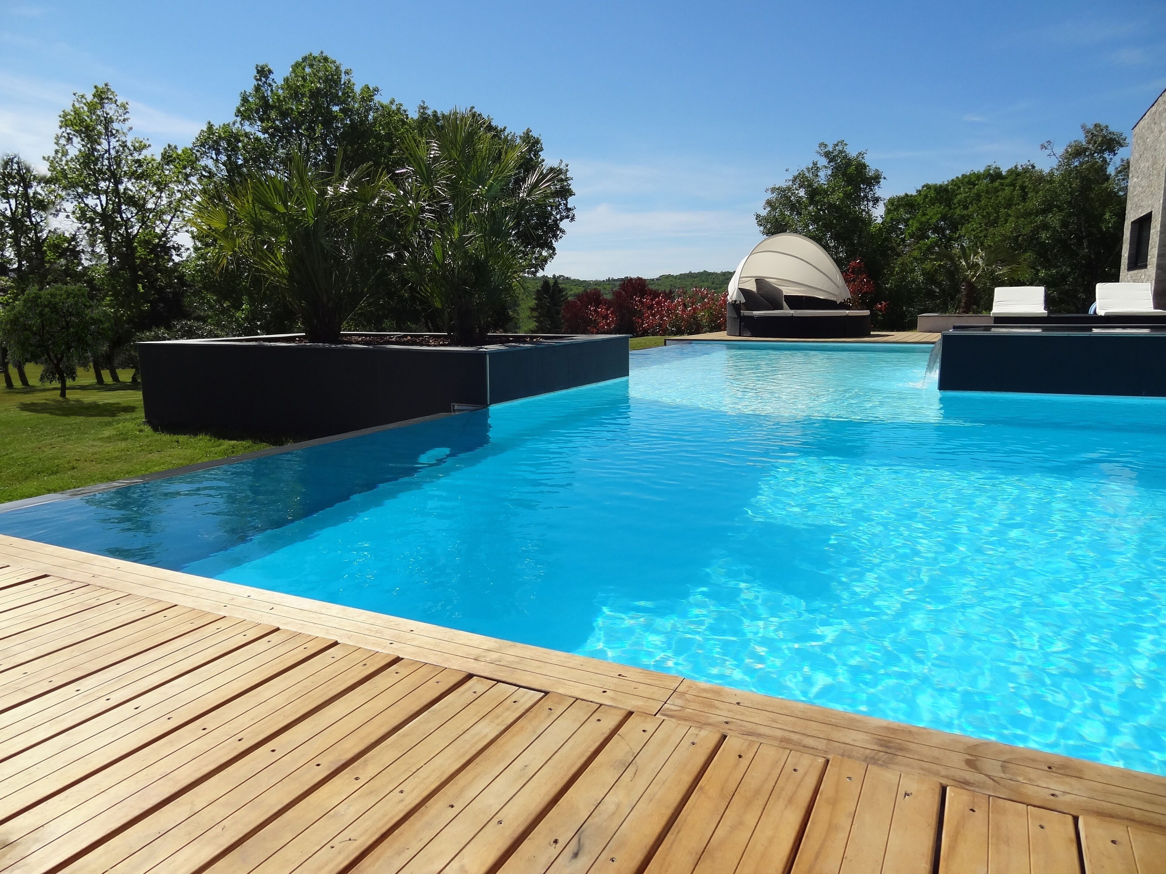 Une piscine dotée d’une terrasse en bois, entourée d’arbres et agrémentée d’un espace couvert avec des chaises longues.