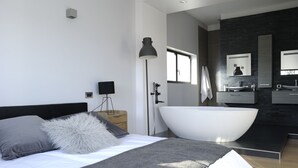 Een moderne slaapkamer met een groot bed, een bedlampje, een nachtkastje en uitzicht op een badkamer met een badkuip en een wastafel.