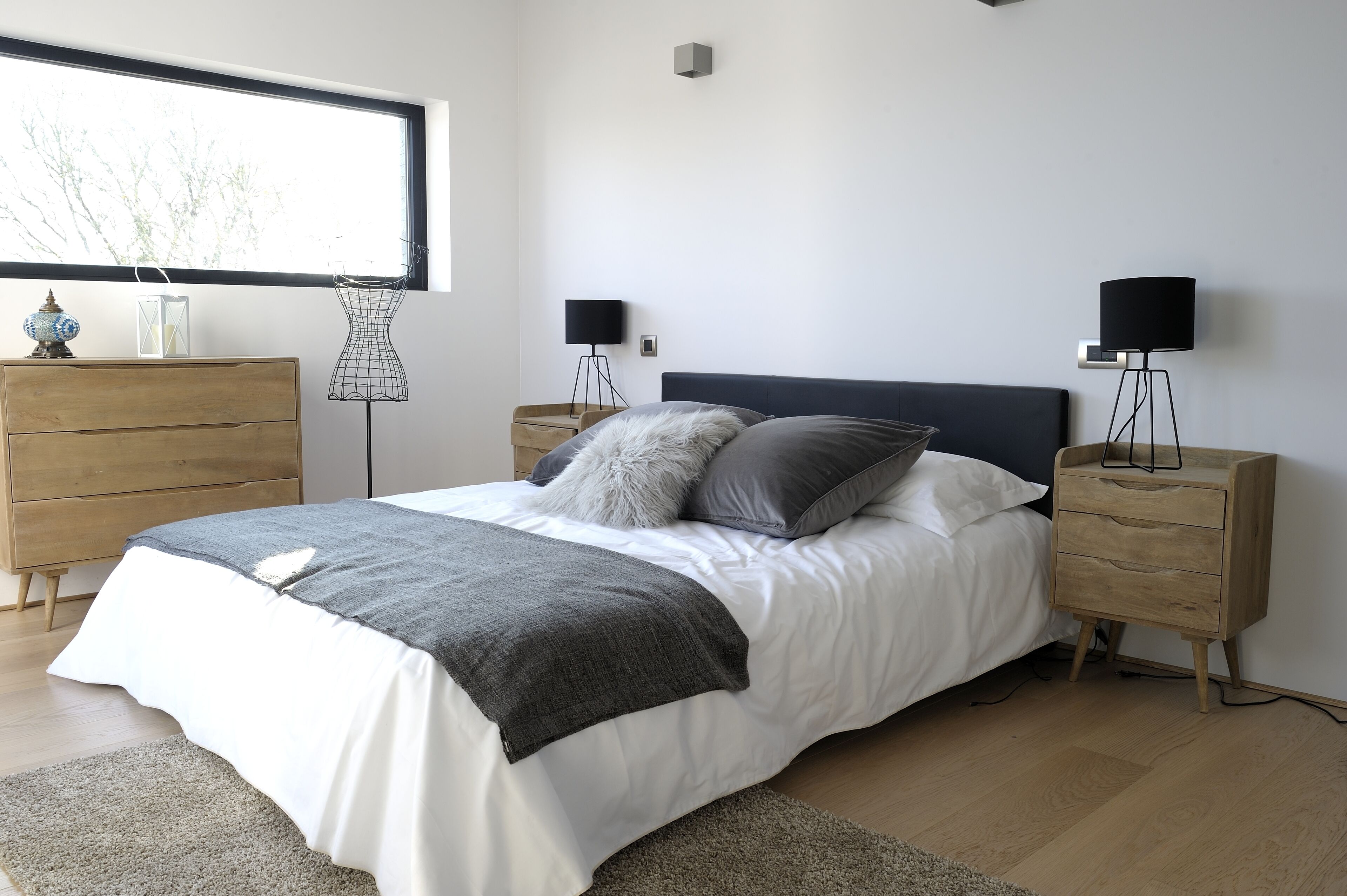 Ein modernes Schlafzimmer mit einem großen Fenster, einer hölzernen Kommode, einem Bett mit weißer und grauer Bettwäsche, zwei Nachttischen mit Lampen und einem kleinen dekorativen Gegenstand auf der Kommode.