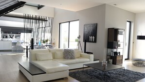 Een moderne woonkamer met een witte bank, een salontafel en een zwart tapijt. Achtergrond toont een keuken met een eethoek.
