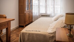 Un dormitorio con un armario de madera, una cama con cubrecama blanco, una mesita de noche de madera y una ventana con cortinas de gasa.
