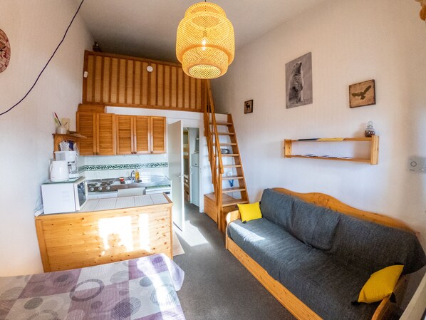 A compact living space with a kitchenette, a wooden loft bedroom, a sofa with cushions, and a hanging light fixture.