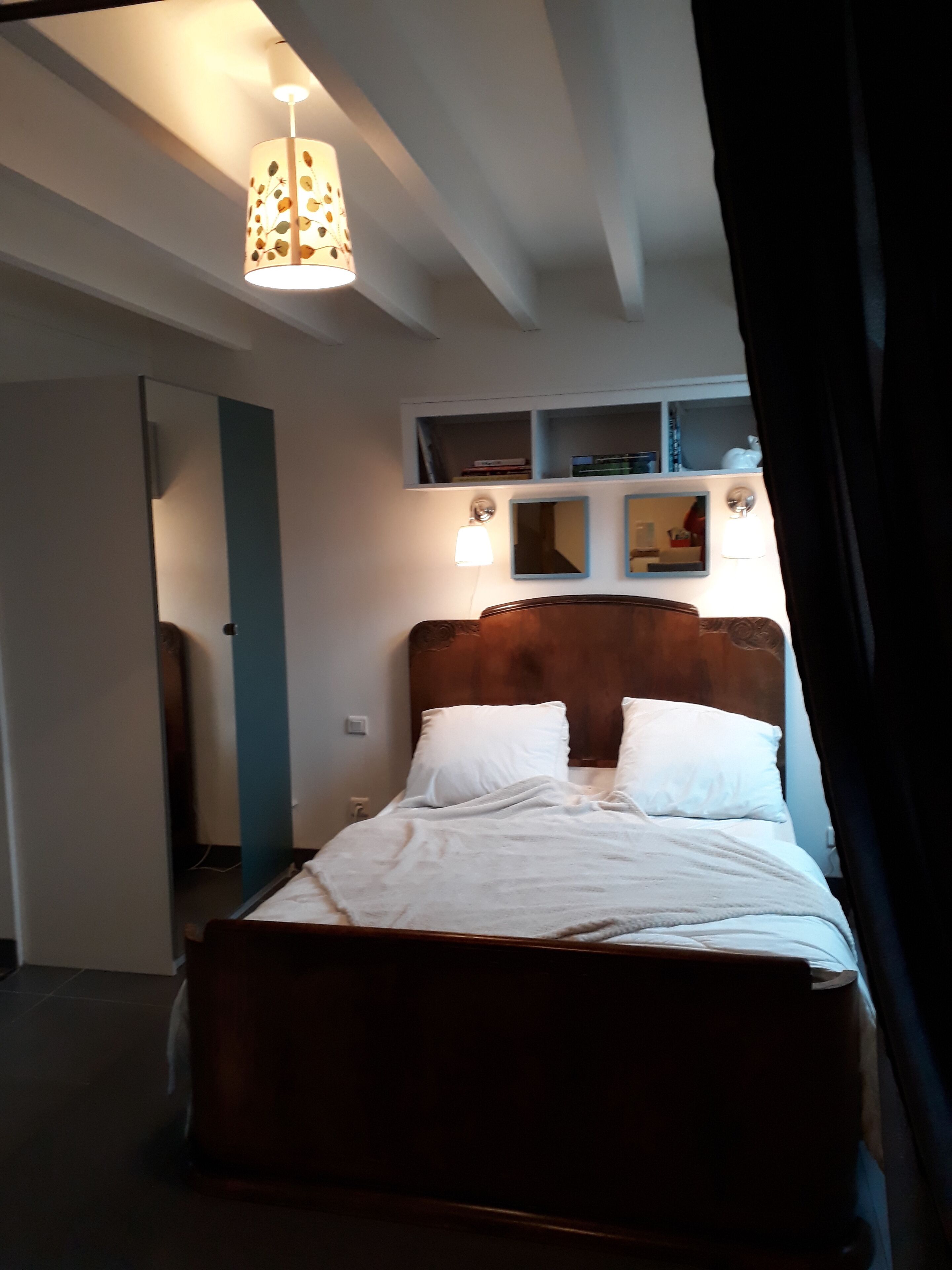 Un dormitorio con una cama de madera, ropa de cama blanca, una lámpara colgante y un estante en la pared con libros.