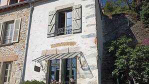 Un edificio de piedra con balcón, una ventana pequeña con contraventanas y una maceta con flores rosas.