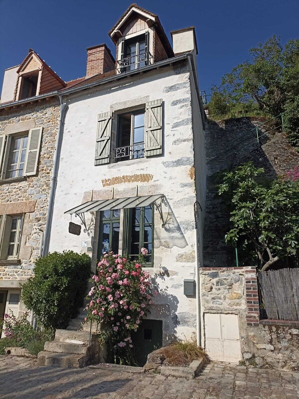Un edificio de piedra con balcón, una ventana pequeña con contraventanas y una maceta con flores rosas.