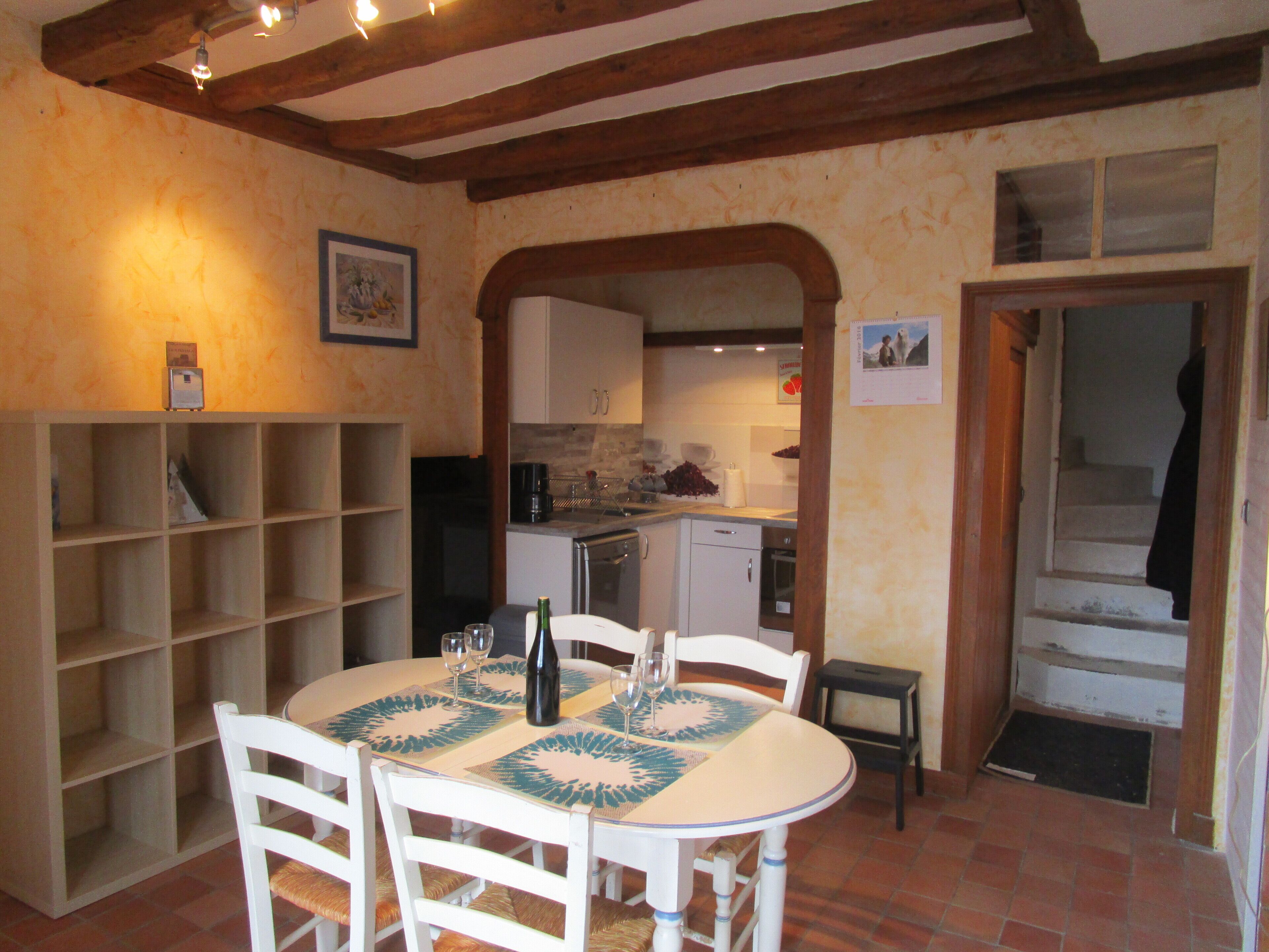 A dining area with a round table, chairs, and a wine bottle. There is a kitchen in the background with cabinets and a sink.