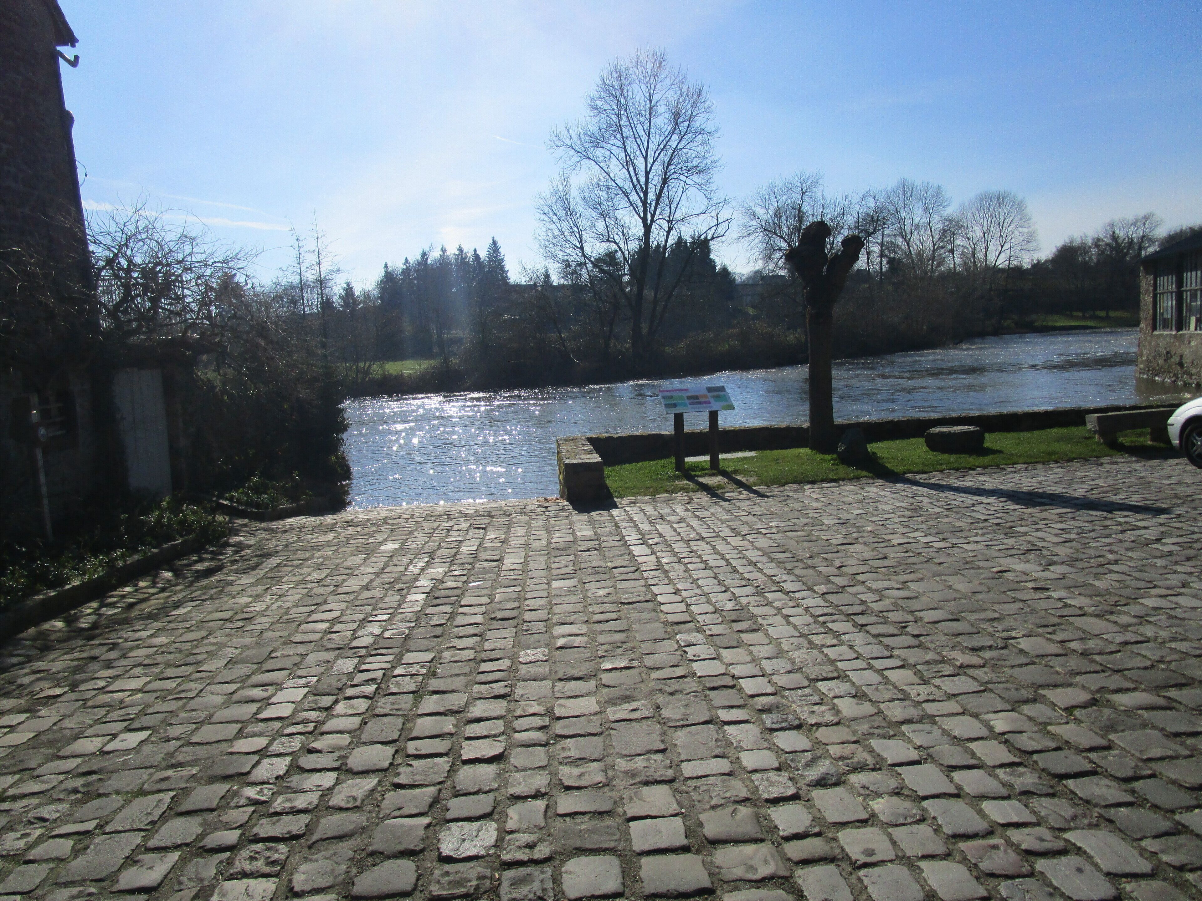 A cobblestone pathway leading to a river with a bench and a signboard.