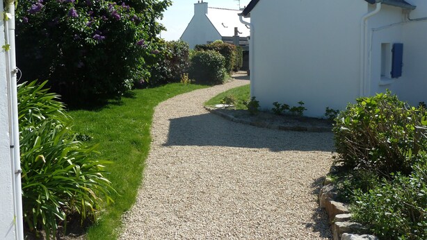 Un chemin de gravier mène à une maison blanche dotée d’un toit en pente, entourée d’une végétation luxuriante et d ’arbustes en fleurs.