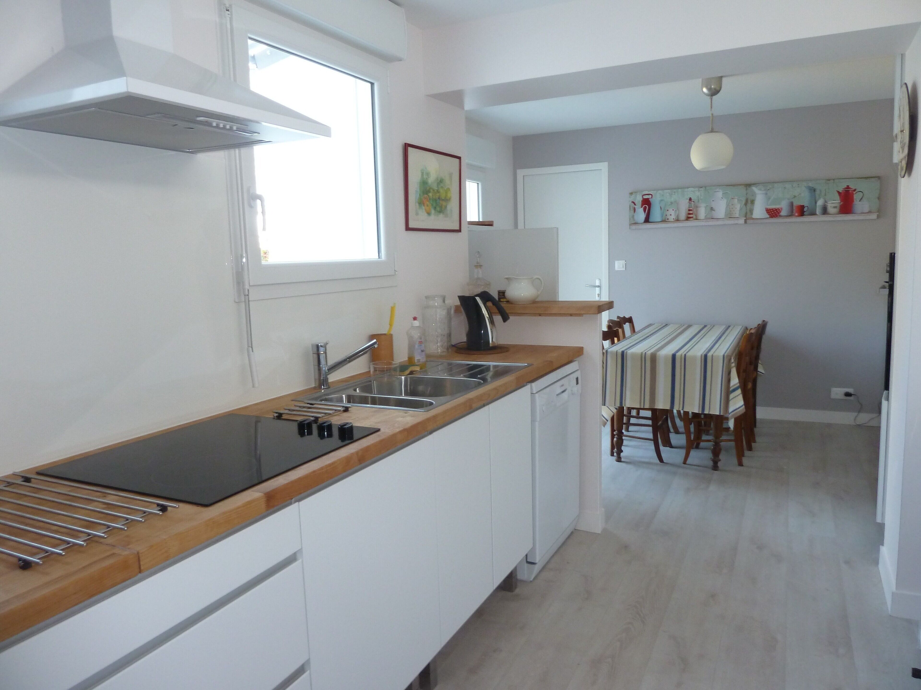 Una cocina moderna con encimera de madera, armarios blancos, lavavajillas integrado y comedor con mantel a rayas y sillas.