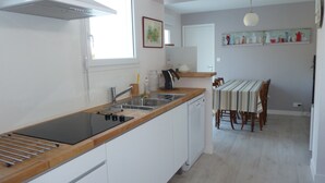 Une cuisine moderne avec un plan de travail en bois, des armoires blanches, un lave-vaisselle encastré et un coin repas agrémenté d’une nappe à rayures et de chaises.