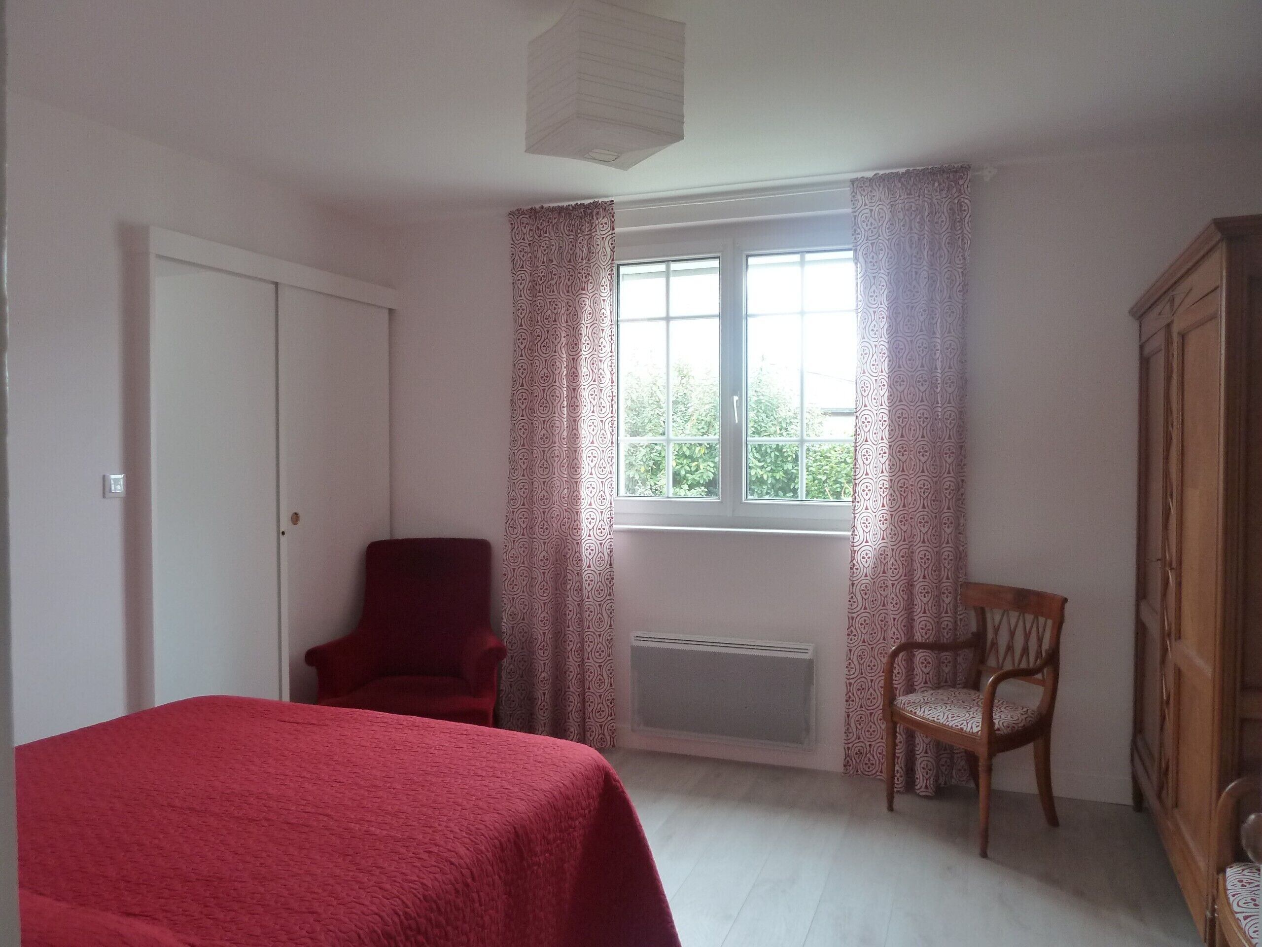 Ein Schlafzimmer mit einem Bett, zwei Stühlen, einem Kleiderschrank, einem Heizkörper und einem Fenster mit Vorhängen.