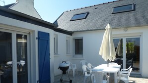 Une terrasse avec un sol en pierre, des chaises en plastique blanches, une table, un barbecue et une porte bleue.