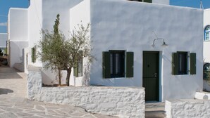 Edifici blanc amb entrada enllosada, un arbre petit i una porta amb persianes verdes.
