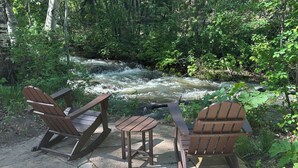 Ein Sitzbereich mit Adirondack-Stühlen und einem kleinen Tisch, mit Blick auf einen Fluss in bewaldeter Landschaft.
