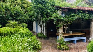 A rustic stone building with a wooden bench and a table, surrounded by lush greenery and a brick pathway.