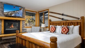 A bedroom with a large bed, wooden headboard, a fireplace, a television mounted above the fireplace, and a window with a view of trees.