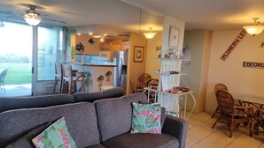 A living room with a grey sofa, two floral cushions, a dining area with wicker chairs, and a kitchen in the background.