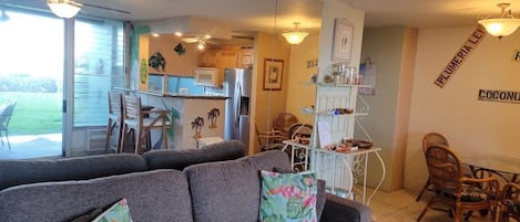 A living room with a grey sofa, two floral cushions, a dining area with wicker chairs, and a kitchen in the background.