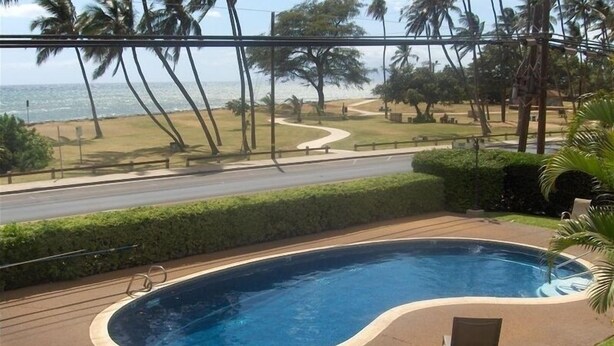 A swimming pool with lounge chairs, surrounded by a well-maintained lawn and palm trees, with a view of the ocean and a road in the background.