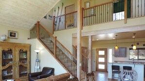 A spacious living area with a high ceiling, a wooden staircase, a dining table set for four, and a kitchen in the background. - New-ish 3 BR house on canyon ridge/Borders Forest/31 windows- 3 miles from town (Pagosa Springs)