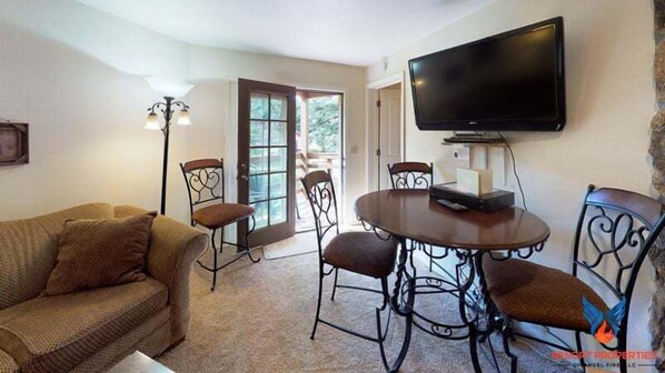 A living room with a sofa, a dining table with chairs, a flat-screen TV mounted on the wall, and a floor lamp.