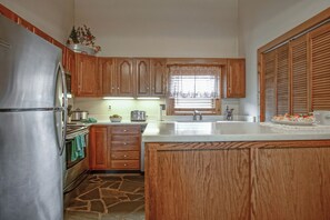 A kitchen with wooden cabinets, a stainless steel refrigerator, a stone backsplash, and a window with a sheer curtain.