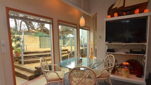 A dining area with a glass table and wicker chairs, a television on a shelf, and a view of a sandy outdoor area through glass doors. - Magnificent Ocean views! On Oceano sandy beach with fire pit  (Oceano,)