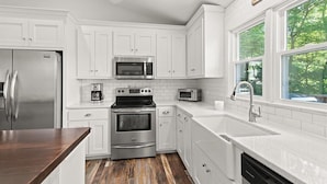 A modern kitchen with white cabinetry, stainless steel appliances, and a large window providing a view of greenery outside. - USAF VETERAN OWNED - DEEP WATER, HUGE HOME, Fire Pit, WiFi, Kayaks/SUP's! (Huddleston)