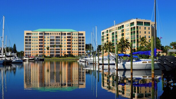 A marina with multiple boats docked, surrounded by buildings with balconies and a clear blue sky.