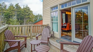 Una terraza de madera con sillas Adirondack, un ventanal que da a un comedor interior y vistas a árboles al fondo.