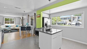 Una cocina moderna con una isla central, un comedor con mesa de vidrio y vistas a autos estacionados afuera.