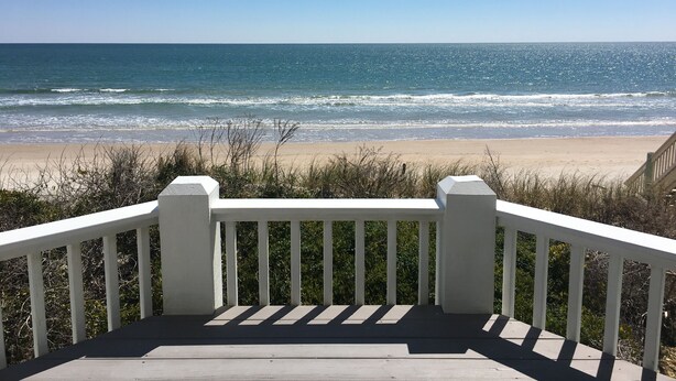 Une terrasse en bois, agrémentée de rampes blanches, qui offre une vue sur une plage où les vagues se brisent sous un ciel bleu et dégagé.