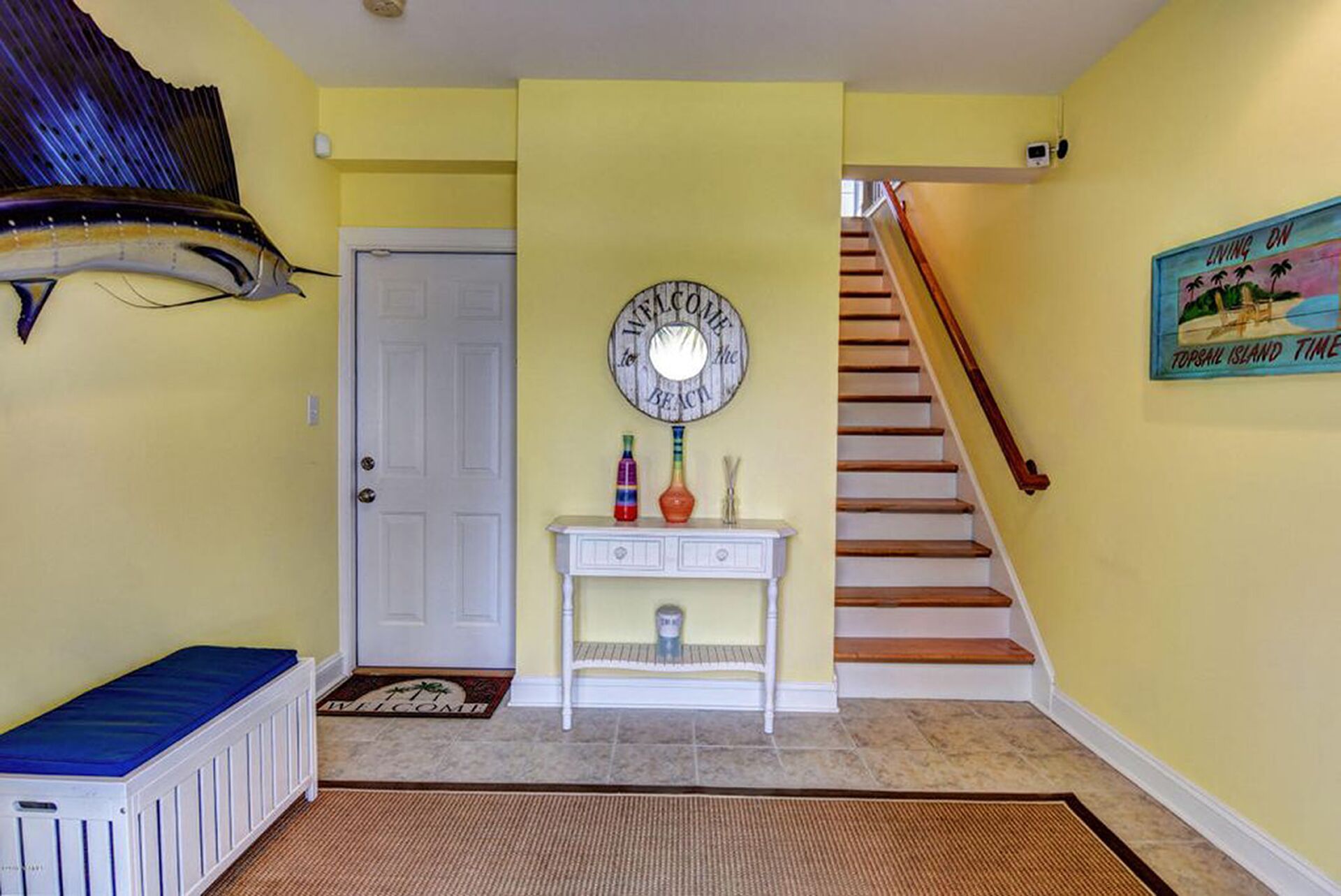 Un couloir avec une porte blanche, un paillasson, une console ornée de bouteilles décoratives, une horloge et un escalier.