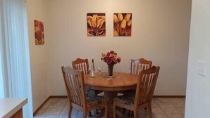 Uma sala de jantar com uma mesa redonda, seis cadeiras e um vaso com flores. Há três obras de arte emolduradas na parede acima da mesa.