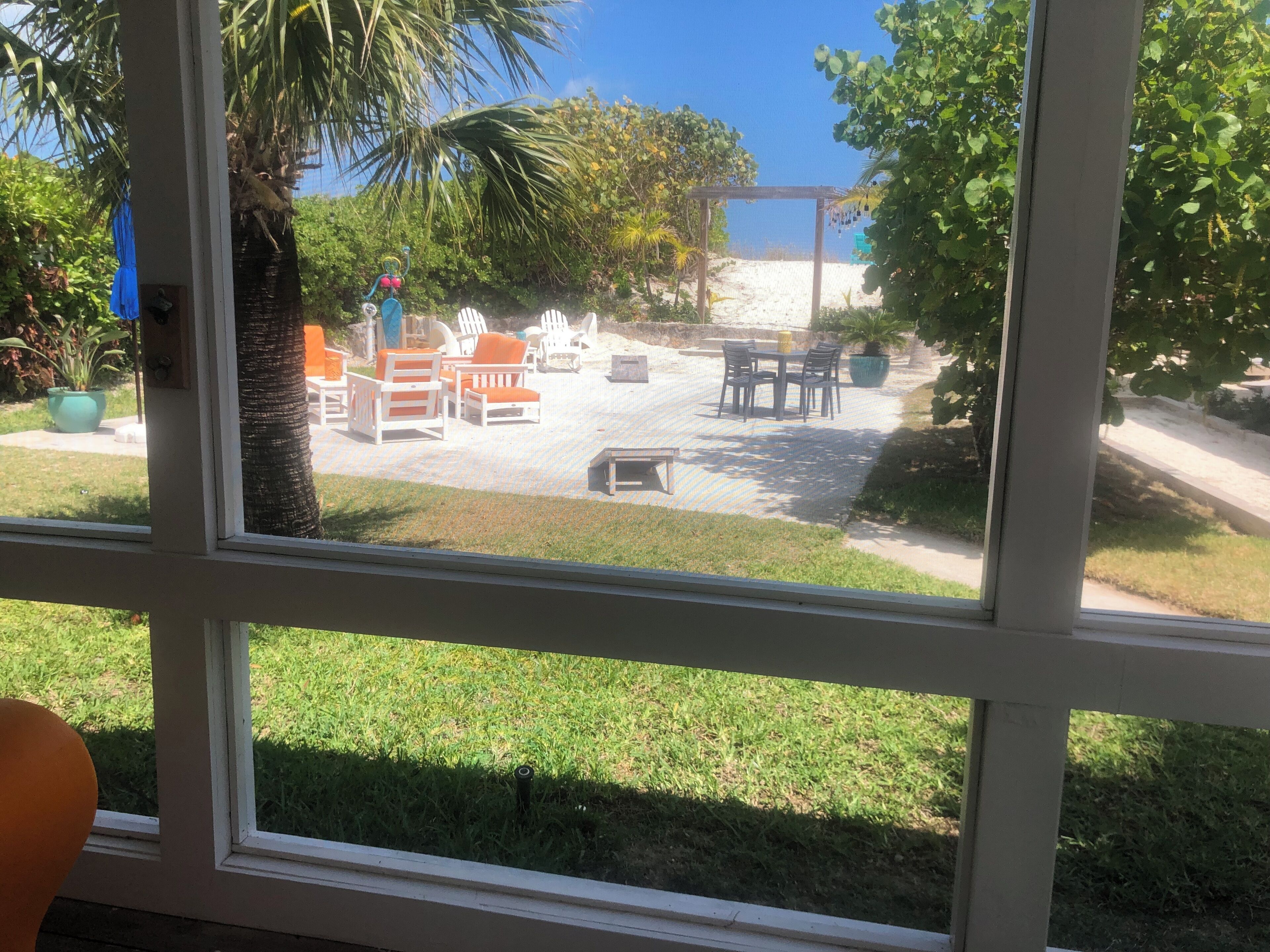 Une vue à travers une fenêtre sur un patio aménagé avec des meubles d’extérieur, une pergola et, au loin, une plage de sable.